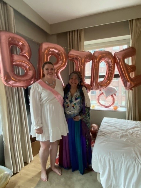 Two women smiling in front of rose gold balloons spelling "BRIDE," with one wearing a white dress and sash, and the other in colorful attire.