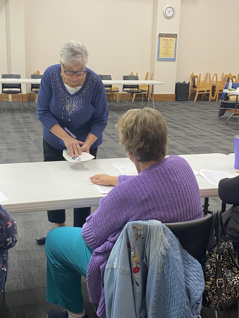 A woman demonstrates a task while others watch at a table in a room with chairs and tables. A clock and framed item hang on the wall.