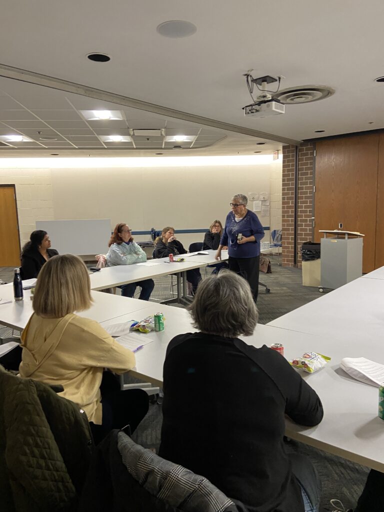 A person stands at the front of a room speaking to a group seated around a large, rectangular table.